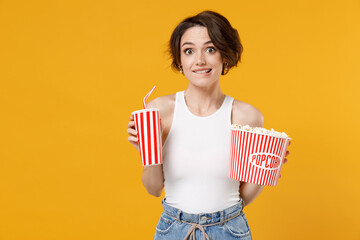 Young fun happy amazed cheerful caucasian surprised woman 20s wearing white tank top shirt holding soda cola popcorn takeaway bucket watching movie film isolated on yellow background studio portrait. © ViDi Studio