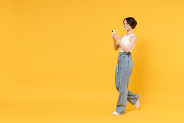 Full length side view young smiling happy woman 20s with bob haircut in white tank top shirt use mobile cell phone chat online browsing surfing internet isolated on yellow background studio portrait