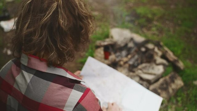 Man with map sits in front of bonfire in the forest.
