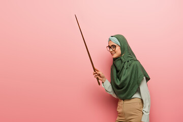 smiling muslim teacher looking away while holding pointing stick on pink background