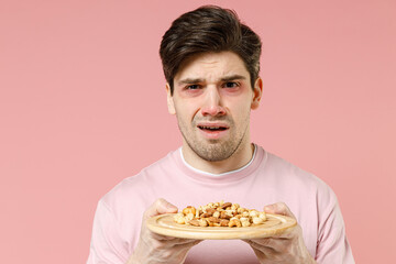 Sick unhealthy ill allergic man has red swelling watery eyes, runny stuffy sore nose suffer from food allergy trigger symptoms feel bad hold plate nuts isolated on pastel pink color background studio.