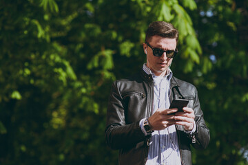 Fototapeta premium Young stylish caucasian man 20s wearing black leather jacket eyeglasses walk in green park in open air using mobile cell phone listen to music in earphones. Concept of urban lifestyle spring season