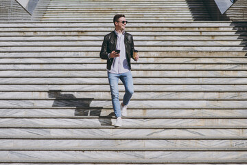 Full length smiling young man in black leather jacket glasses walk in city downtown go down concrete steps hold mobile cell phone takeaway craft paper cup drink coffee to go Urban lifestyle concept.