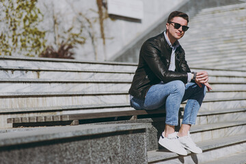 Full length young stylish fun smiling happy man 20s wear black leather jacket eyeglasses sitting on city concrete steps, resting in free time, walk in downtown, look aside Concept of urban lifestyle
