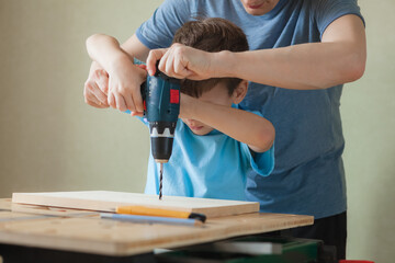 Dad and son work together while standing at workbench. Little boy learning to drill. Cute child holding drill screwdriver and man helping him