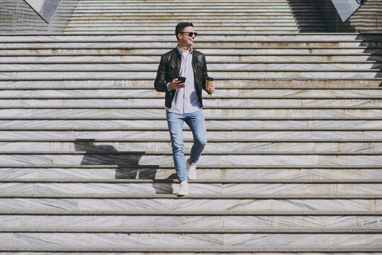 Full length smiling young man in black leather jacket glasses walk in city downtown go down concrete steps hold mobile cell phone takeaway craft paper cup drink coffee to go Urban lifestyle concept. - Powered by Adobe