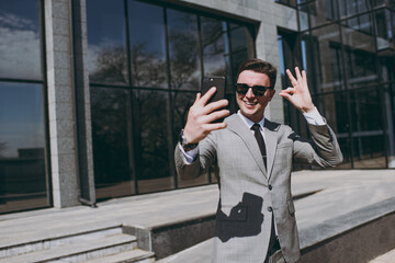 Young successful employee business man in grey suit walk go near office glass wall building outdoors in downtown city center doing selfie shot on mobile phone for social network show ok okay gesture.