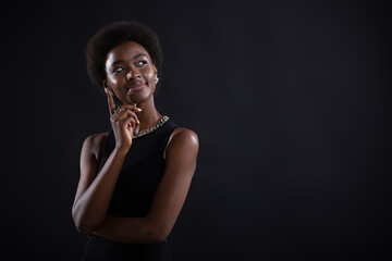 Young black woman trying to find best solution and searching answer. African american female having doubtful expression looking side away and holding her chin with hand on black background.