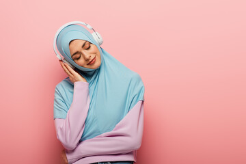 Obraz premium pleased muslim woman listening music in headphones with closed eyes on pink background