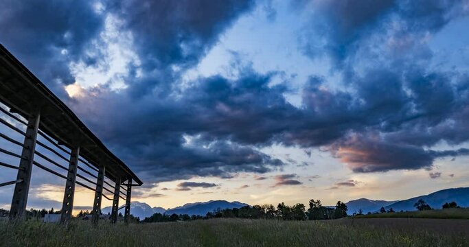Time lapse of beautiful and dramatic sunset over grassland farming field and Alps mountains in the distance. Wooden hayrack decorating the field in Slovenia. Wide angle, Zoom in