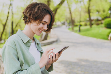 Young side view smiling student happy woman in casual jacket sit on bench outdoors in city spring park blur background use mobile cell phone chat online in social network. People lifestyle concept