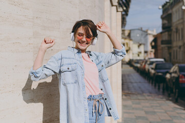 Young happy fun smiling caucasian woman 20s wearing jeans clothes headphones eyeglasses listening...