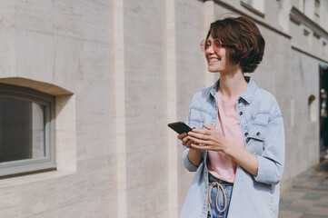 Young smiling student woman in jeans clothes eyeglasses walk down city street near building wall outdoors hold mobile cell phone using online internet maps look aside People urban lifestyle concept