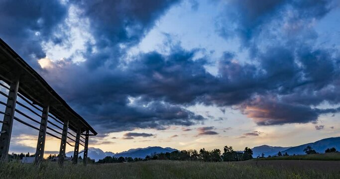 Time lapse of beautiful and dramatic sunset over grassland farming field and Alps mountains in the distance. Wooden hayrack decorating the field in Slovenia. Wide angle, right trucking