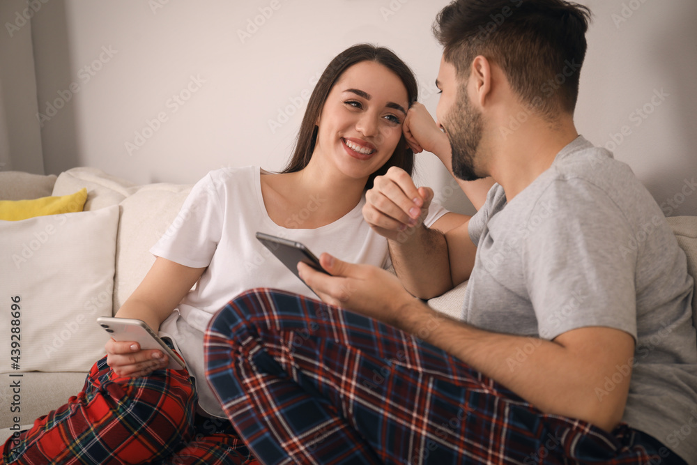 Poster Happy couple in pajamas with gadgets on sofa at home