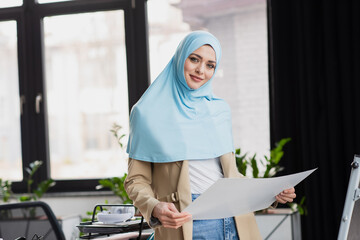 Fototapeta na wymiar pretty muslim businesswoman looking at camera while holding paper in office