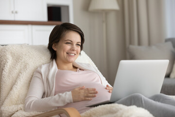 Smiling pregnant woman using laptop, looking at screen, touching caressing belly, sitting in cozy chair at home, happy young future mom watching movie or making video call, shopping online