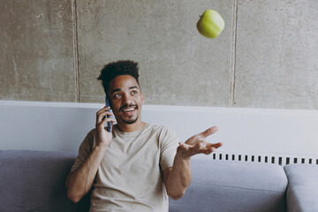 Happy vegeterian young african american man in beige t-shirt sit on grey sofa indoors apartment talk by mobile cell phone with friends toss up apple rest on weekends stay home Tattoo translate fun