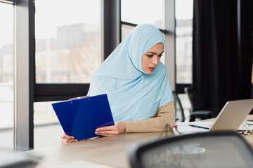 pretty muslim businesswoman holding clipboard while looking at laptop, blurred foreground