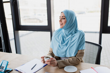 smiling muslim businesswoman with cup of coffee sitting at workplace against window
