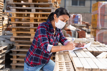Hispanic woman supervisor in protective face mask controlling quantity of redbricks at hardware store warehouse