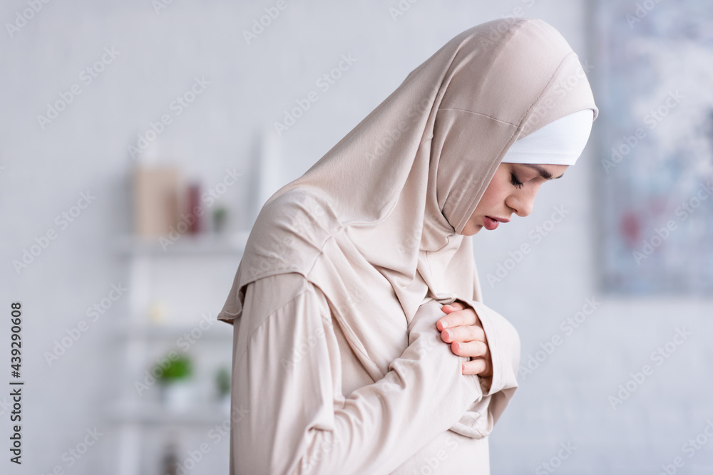 Wall mural young muslim woman holding hands on chest while praying with closed eyes