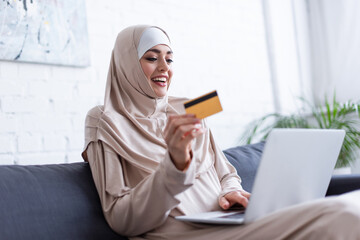 smiling muslim woman holding digital tablet near blurred laptop at home