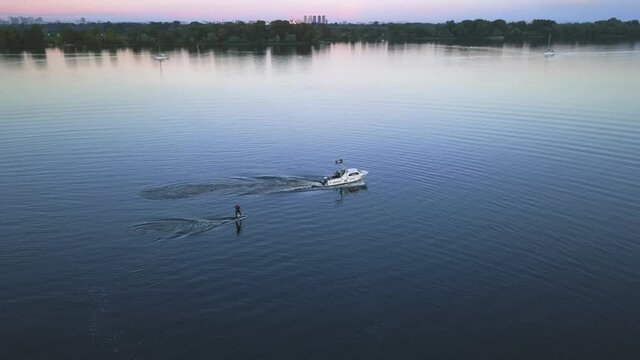 The athlete gets up from the water on foilboarding holding on to the rope tied to the boat in the evening aerial view