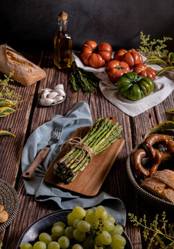 Dark photography of asparagus, bread, tomatoes,garlic and plums on a table of old wood. Vegetables recipe of for autumn or winter. Moody top view of seasonal products on a wooden table.