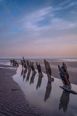 Reflections of wooden groynes on a beach with the ocean in the distance under a sunset sky.