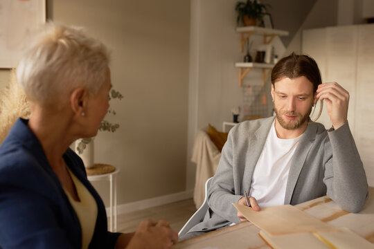 Middle Aged Blonde Female Mentor Or Coach Talking To Young Male, Teaching Intern Having Business Conversation With Workmate. Mature Woman Helping Man Colleague, Discussing New Project In Office