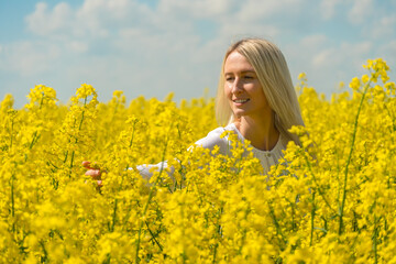Naklejka premium Smiling young blonde woman with her arm outstretched in a white shirt in a rapeseed field touching flowers on a sunny day