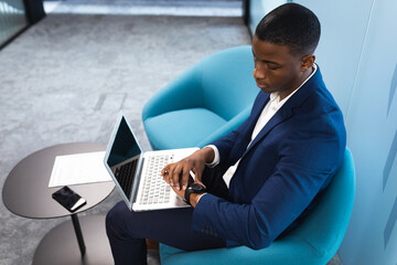 African american businessman with laptop using smartwatch while sitting on a chair at modern office