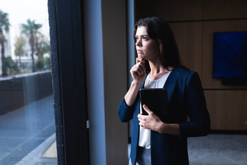 Thoughtful caucasian businesswoman holding digital tablet looking out of window at modern office