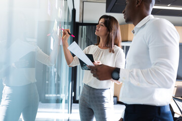 Diverse businessman and businesswoman writing on memo notes on glass board at modern office