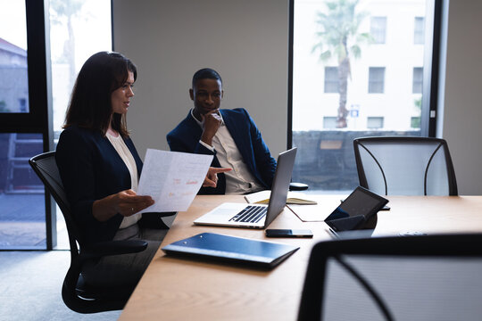 Diverse businessman and businesswoman discussing over laptop in meeting room at modern office