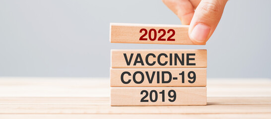 hand pulling 2022 block over Vaccine, COVID-19 and 2019 wooden building on table background. Crisis...