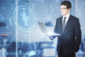 Attractive young european businessman with laptop standing in office with creative glowing blue business interface on blurry background. Human resources and online recruitment concept