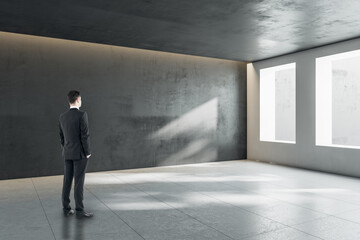 Thoughtful young businessman standing in concrete interior with mock up place and windows with daylight.