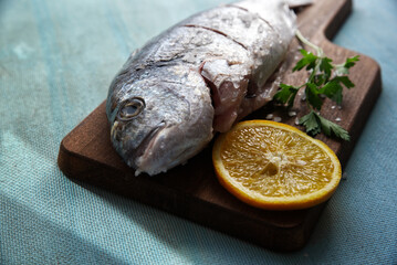 Fish with orange slices and parsley, on a wooden board and a light blue table in the background.