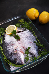 Fish in a glass tray with coarse salt, lemon slices and parsley on a dark background.