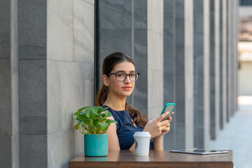 Young attractive girl holding  a smart phone checking or making social connection at an outdoor café on the street. Woman in casual using technology while having tea or coffee outside