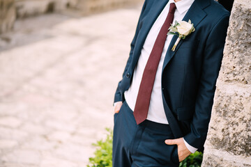 The groom in the suit with a red tie and a boutonniere with white rose on the street of the old town, close-up - obrazy, fototapety, plakaty
