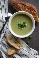 Green peas soup in a white bowl, fresh baguette bread on striped kitchen towel. Vegetarian meal top view photo. Eating fresh concept.  