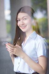 Portrait of an adult Thai student in university uniform.  Asian beautiful woman standing near the window happily smiling uses her smartphone to search for educational information.