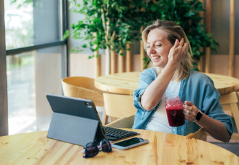 Young happy cheerful laughing blonde working on a laptop sitting at a table in a cozy cafe and...