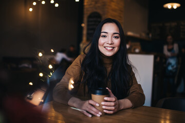 Smiling young Asian lady drinking hot coffee in cafeteria