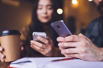 Cropped image of male and female hands using modern cellphone devices for online communication during organisation planning in coffee shop, selective focus on digital smartphones and takeaway cup
