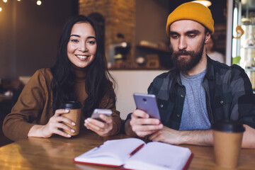 Positive couple using smartphone while sitting at table in cafe and drinking coffee