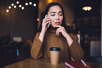 Pensive female student using smartphone gadget for making cellphone consultancy conversation during coffee time in cafe interior, contemplative hipster girl pondering on received mobile information